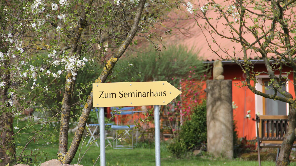 Gelbes Richtungsschild mit der Aufschrift „Zum Seminarhaus“ auf zwei Metallpfosten, eingebettet in eine grüne Gartenlandschaft mit blühenden Bäumen und Gartenmöbeln.