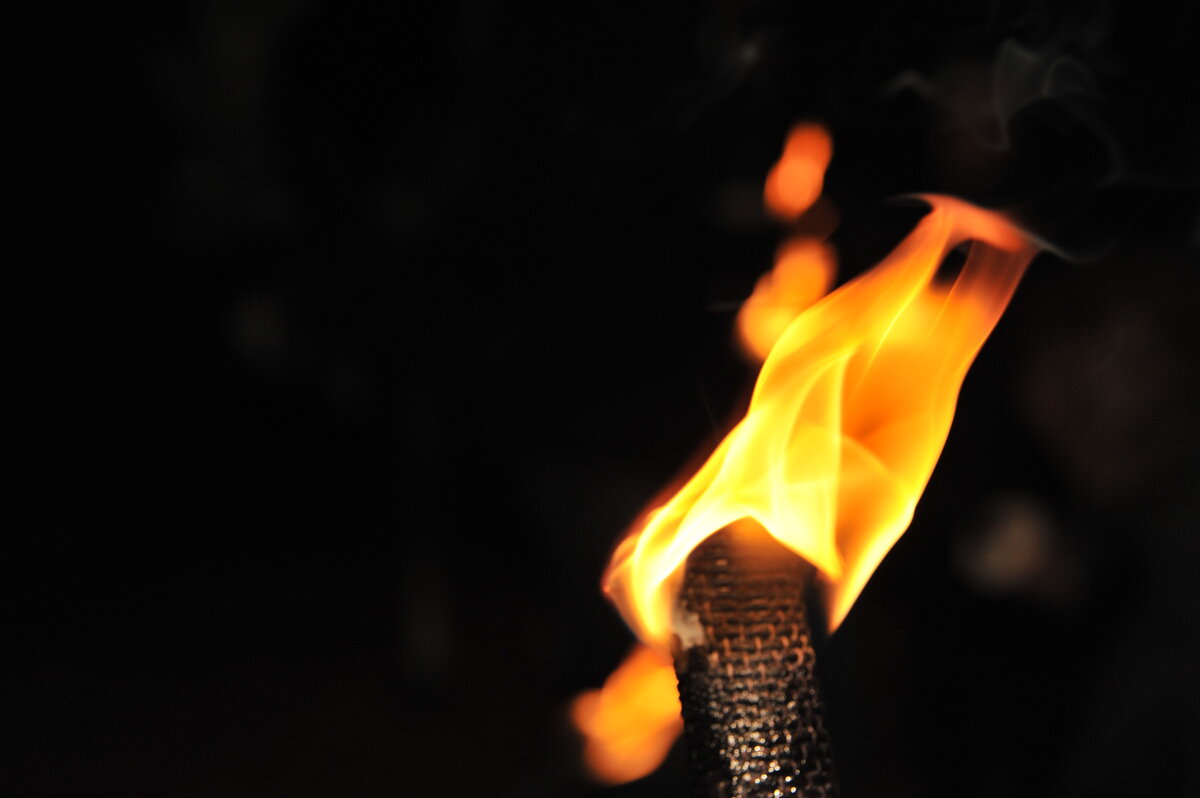 Nahaufnahme einer brennenden Fackel mit leuchtender Flamme vor dunklem Hintergrund – weitere Flammen im Bild erzeugen eine atmosphärische Szenerie.