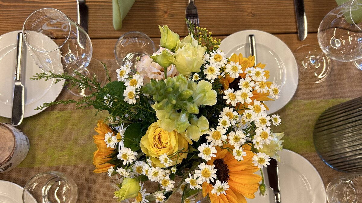Holztisch mit Juteläufer, buntem Blumenarrangement und grünen Stoffservietten für ein festliches Dinner.
