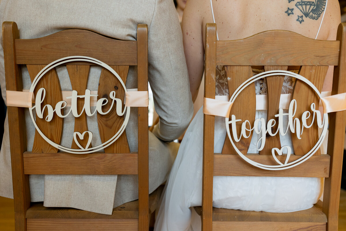 Zwei Holzstühle mit liebevoll gestalteten Schildern: „better“ und „together“ – jeweils mit Herzsymbol und rosa Schleifen befestigt. Die sitzenden Personen tragen ein graues Hochzeitsoutfit und ein weißes Spitzenkleid.