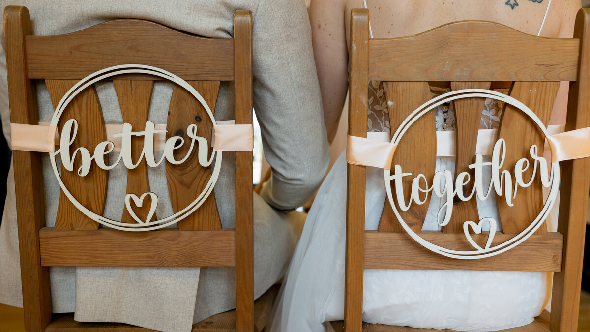 Zwei Holzstühle mit liebevoll gestalteten Schildern: „better“ und „together“ – jeweils mit Herzsymbol und rosa Schleifen befestigt. Die sitzenden Personen tragen ein graues Hochzeitsoutfit und ein weißes Spitzenkleid.