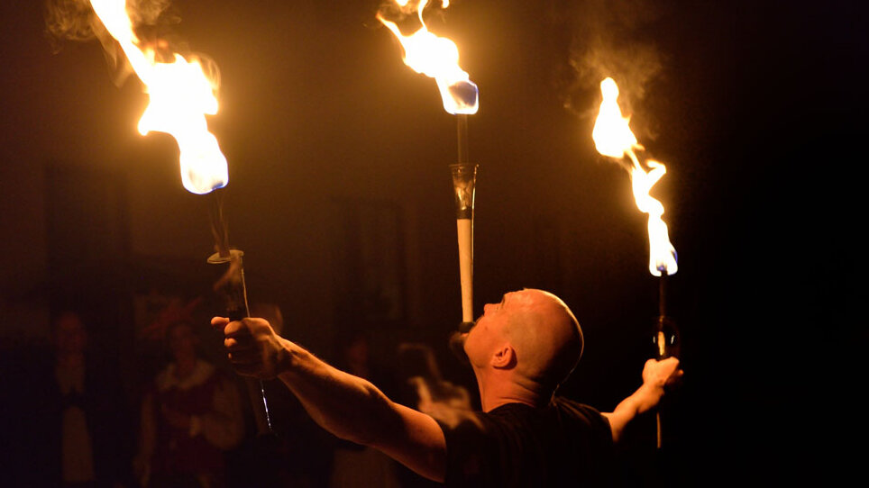 Feuershow bei Nacht mit Artist, der drei brennende Fackeln balanciert und von Publikum beobachtet wird.