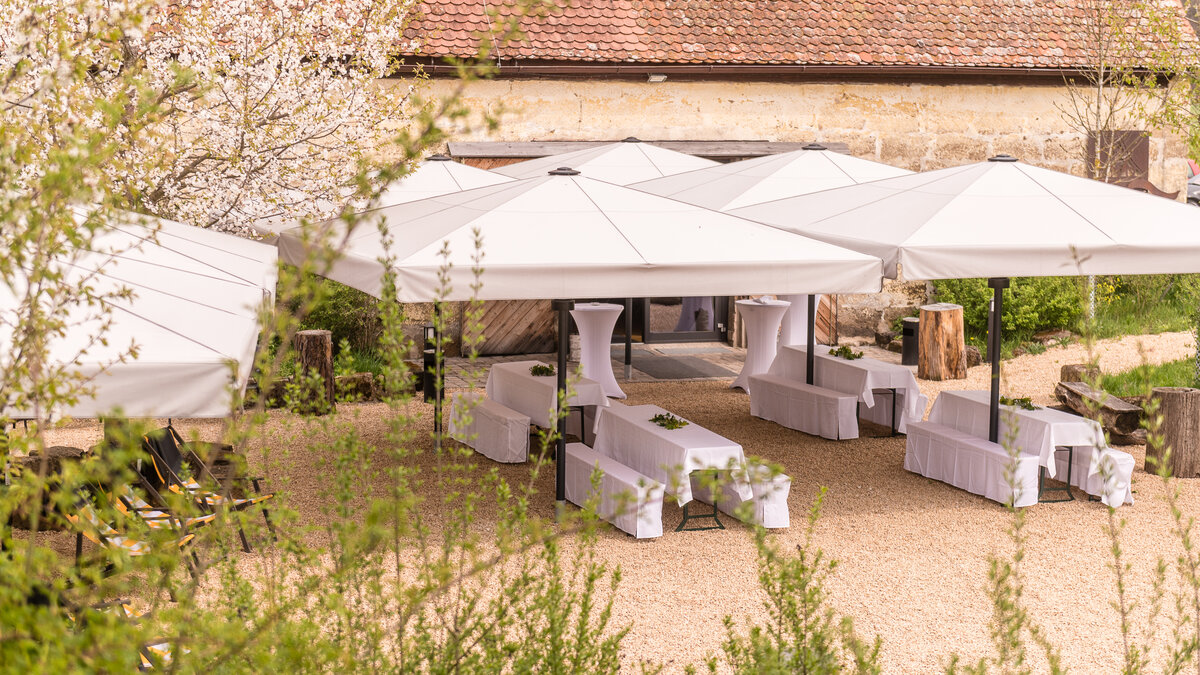 Gartenfest mit weißen Sonnenschirmen, langen Tafeln und blühenden Bäumen im Innenhof.