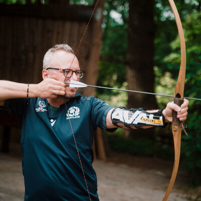 Person beim Bogenschießen mit traditionellem Recurvebogen im Freien, gekleidet in ein „SCOTLAND WORLD CUP“-Shirt und ausgestattet mit Armschutz. Im Hintergrund Bäume und Holzstruktur.