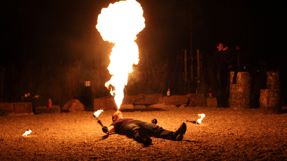 Mann liegt auf dem Boden und spuckt einen riesigen Feuerball in die dunkle Nacht bei einer Feuershow, Flammenwerkzeug liegt neben ihm.