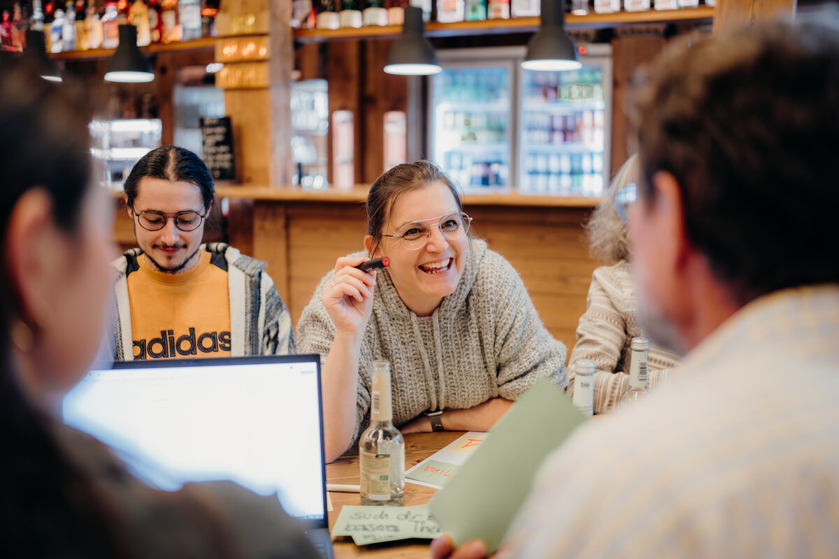 Gruppe von Menschen bei informellem Treffen in gemütlicher Bar mit Laptop, Notizen und Getränken.