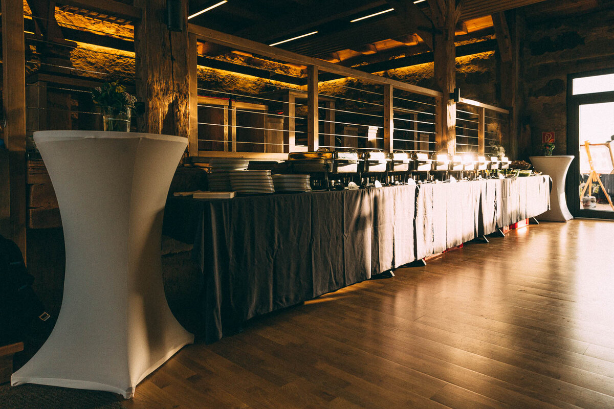 Buffetaufbau in rustikalem Veranstaltungsraum: Graue Tischdecke, Warmhaltebehälter, Tellerstapel und Besteck deuten auf ein festliches Essen hin. Stehtische mit Blumenarrangements runden das gemütlich-elegante Ambiente ab – ideal für Hochzeit, Firmenfeier oder Bankett.