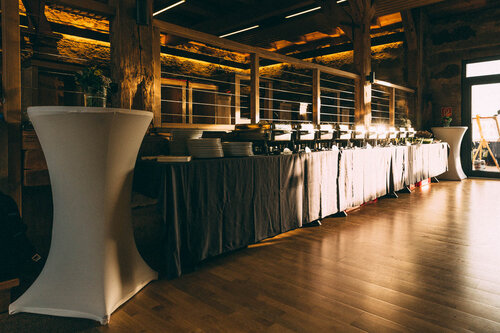 Buffetaufbau in rustikalem Veranstaltungsraum: Graue Tischdecke, Warmhaltebehälter, Tellerstapel und Besteck deuten auf ein festliches Essen hin. Stehtische mit Blumenarrangements runden das gemütlich-elegante Ambiente ab – ideal für Hochzeit, Firmenfeier oder Bankett.