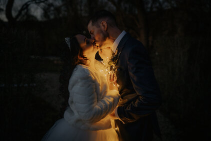  Brautpaar küsst sich bei Nacht, umgeben von warm leuchtender Lichterkette, Braut mit Tiara und Fellschal.