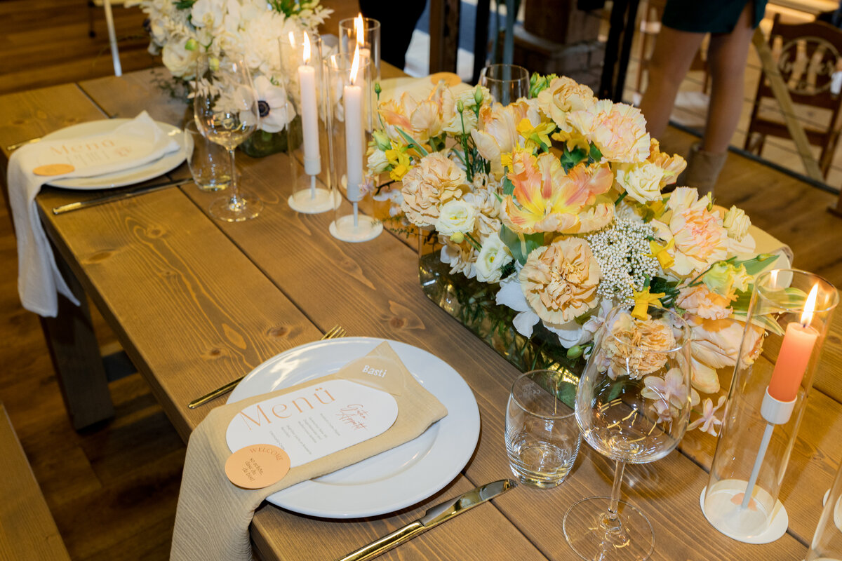 Festlich gedeckter Holztisch mit Menükarte „Basti & Mira“, Blumenarrangement und Kerzen für eine Hochzeit.