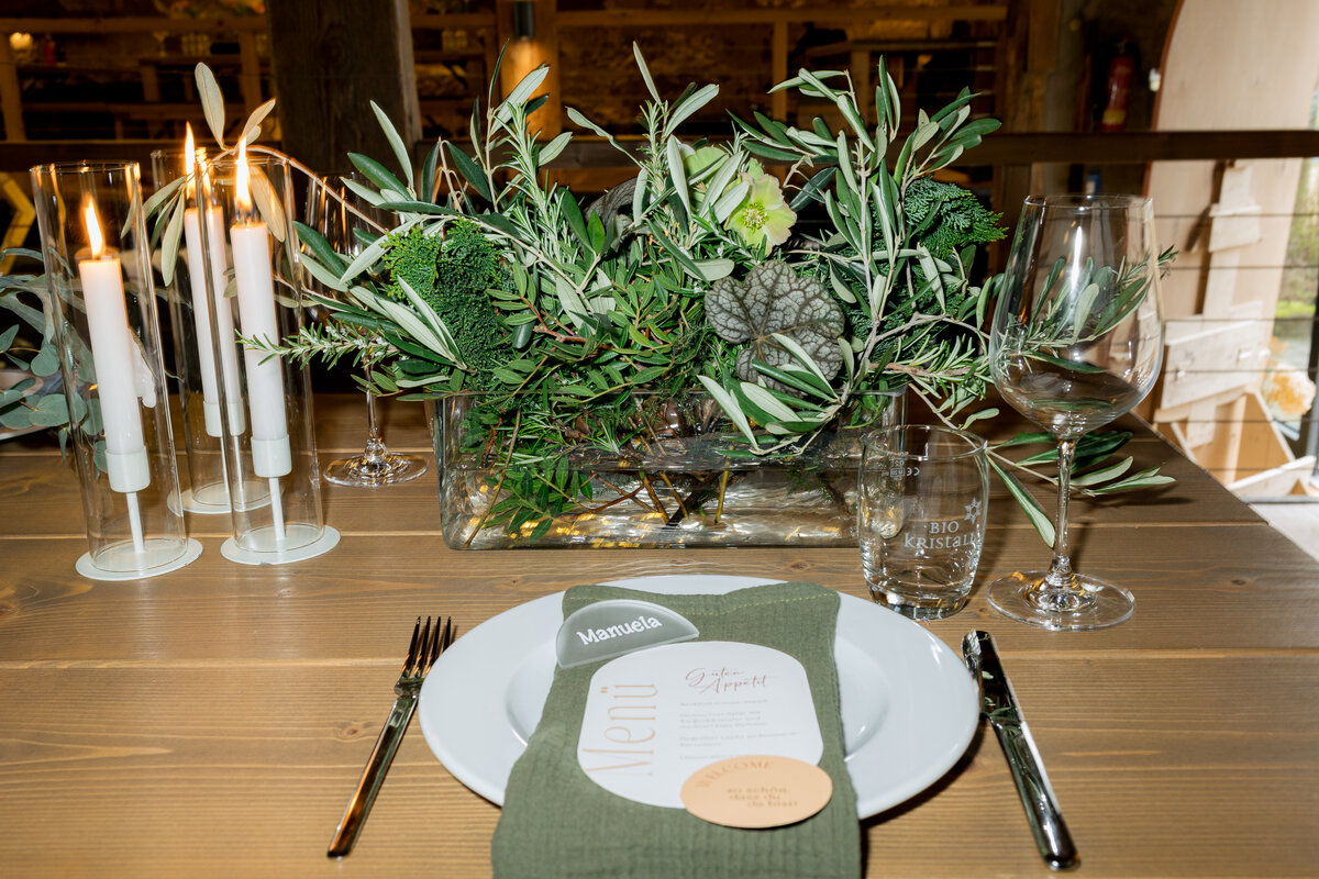  Elegant gedeckter Tisch mit weißem Teller, grünem Stoffserviette, Menükarte „Manuela“, Apfelkuchendessert, Kerzen und Glasvase mit Olivenzweigen als festlicher Mittelpunkt.