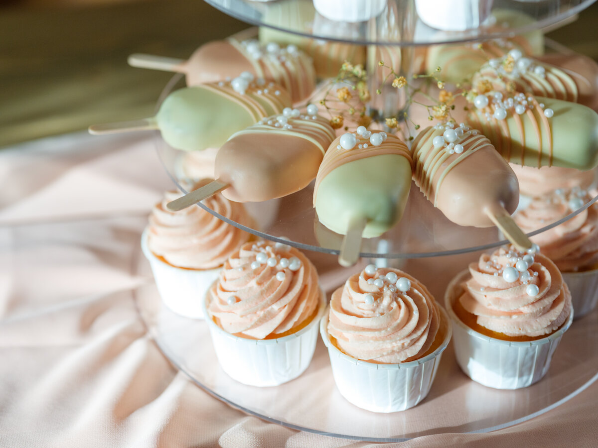 Zweistöckiger Dessertständer mit verzierten Cake Pops und Cupcakes in Pastellfarben auf rosa Stoff.