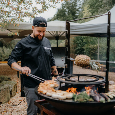 Ein Koch grillt Fleisch und Gemüse auf einem offenen Feuergrill bei einem Outdoor-Event.