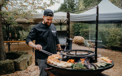 Ein Koch grillt Fleisch und Gemüse auf einem offenen Feuergrill bei einem Outdoor-Event.
